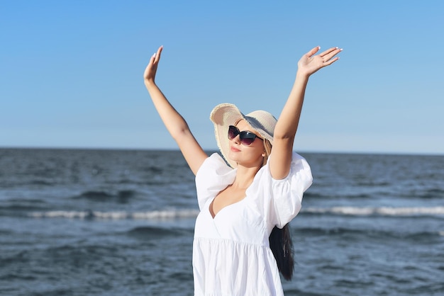 Une femme blonde heureuse est sur la plage de l'océan dans une robe blanche et des lunettes de soleil levant les mains