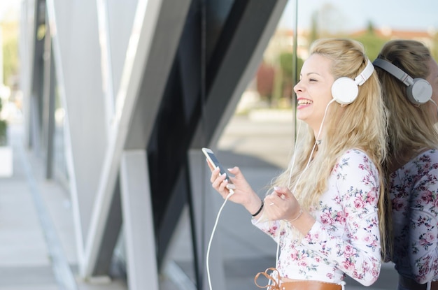 Une femme blonde heureuse isolée écoutant de la musique avec des écouteurs et un téléphone portable à l'extérieur