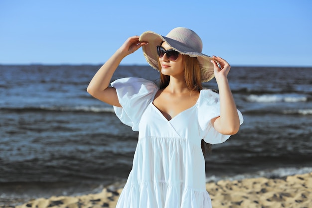 Une femme blonde heureuse pose sur la plage de l'océan avec des lunettes de soleil et un chapeau. Soleil couchant.