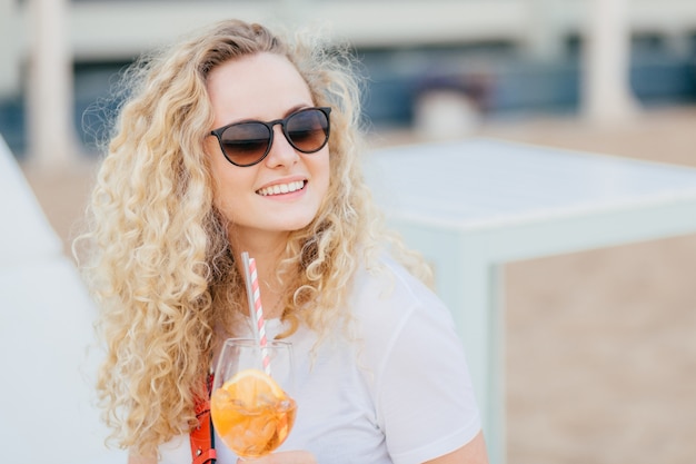 Femme blonde joyeuse aux cheveux clairs et ondulés, sourire positif, porte des nuances, regarde de côté avec une expression heureuse, boit un cocktail frais