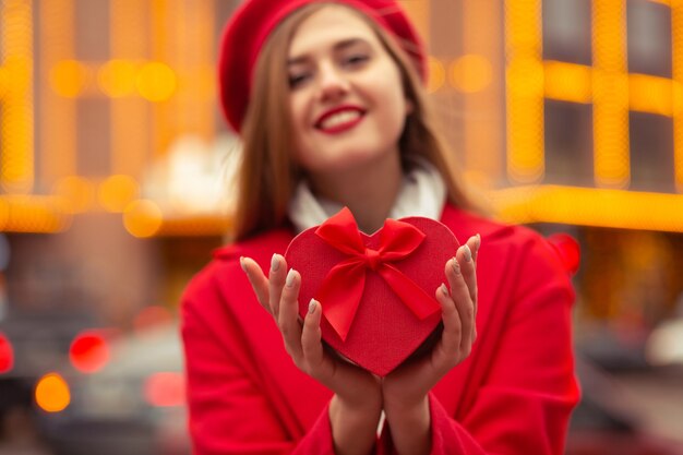 Une femme blonde joyeuse porte un béret rouge et un manteau tenant une boîte-cadeau en forme de coeur à l'arrière-plan des lumières bokeh. Espace pour le texte