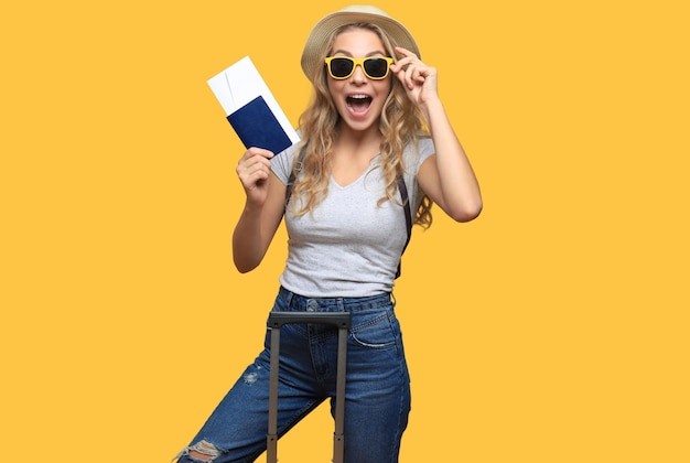 Une femme blonde joyeuse posant avec des bagages et tenant un passeport avec des billets tout en détournant le regard sur un fond jaune