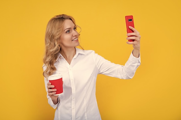 Femme blonde joyeuse avec une tasse de café faisant selfie au téléphone sur fond jaune