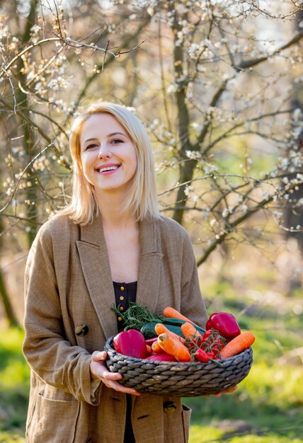 Femme blonde avec des légumes dans un panier dans un jardin fleuri