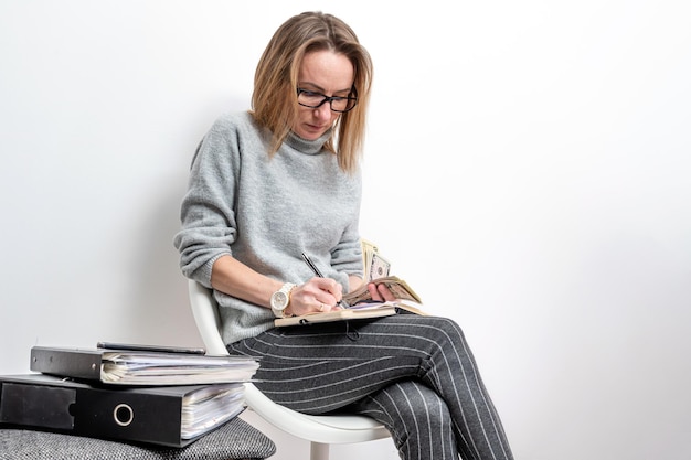 Femme blonde avec des lunettes comptant de l'argent et écrivant dans un cahier isolé sur fond gris