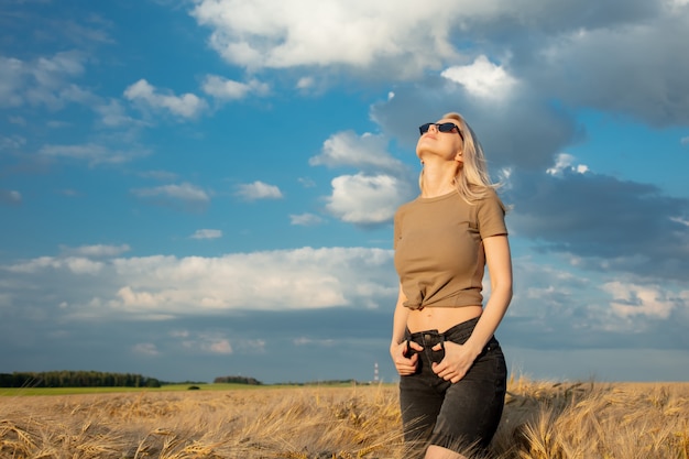 Femme blonde à lunettes de soleil reste dans le champ de blé au coucher du soleil