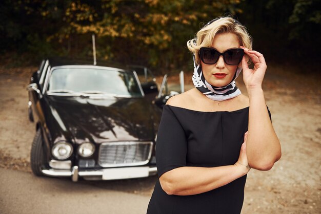 Femme blonde à lunettes de soleil et en robe noire près de la vieille voiture classique vintage.