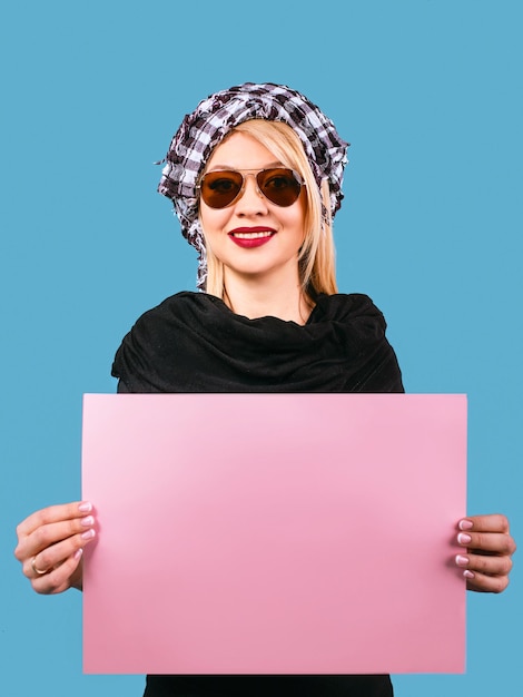 femme blonde à lunettes de soleil et shemag arafatka avec table vide rose