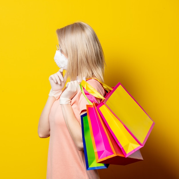 Femme blonde en masque facial avec des sacs à provisions sur mur jaune