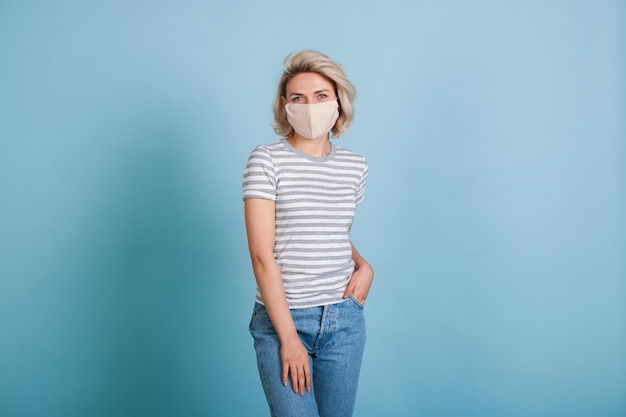 Femme blonde avec masque médical portant des jeans et t-shirt pose sur un mur de studio bleu