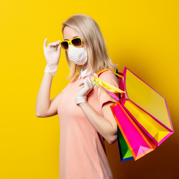 Femme blonde en masque et robe rose détiennent des sacs à provisions sur mur jaune