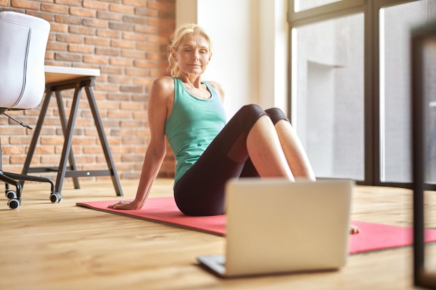 Femme blonde mature ciblée en tenue de sport assise sur le sol et regardant un cours de yoga vidéo en ligne