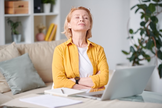 Femme blonde mature sereine dans des vêtements décontractés de détente sur le canapé en face de l'ordinateur portable avec les yeux fermés pendant le travail à distance à la maison