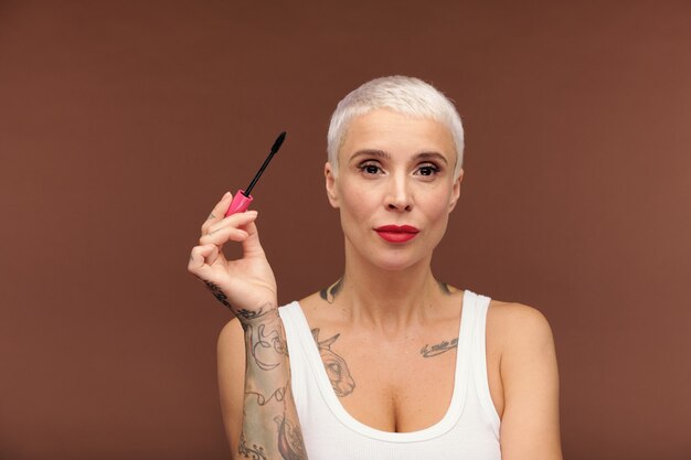 Femme blonde mature avec des tatouages sur les bras et la poitrine et du rouge à lèvres rouge sur les lèvres tenant du mascara noir tout en l'appliquant sur les cils