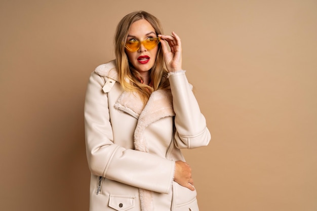 Femme blonde à la mode élégante posant sur fond beige, style de mode hiver automne automne, lunettes de soleil rétro jaunes, maquillage lumineux, vacances d'hiver de luxe, couleurs beiges.