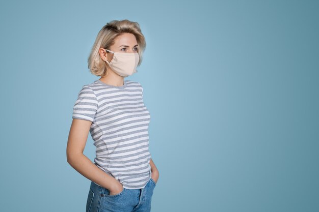 Femme blonde à la mode avec masque médical sur le visage posant sur un mur bleu avec espace libre