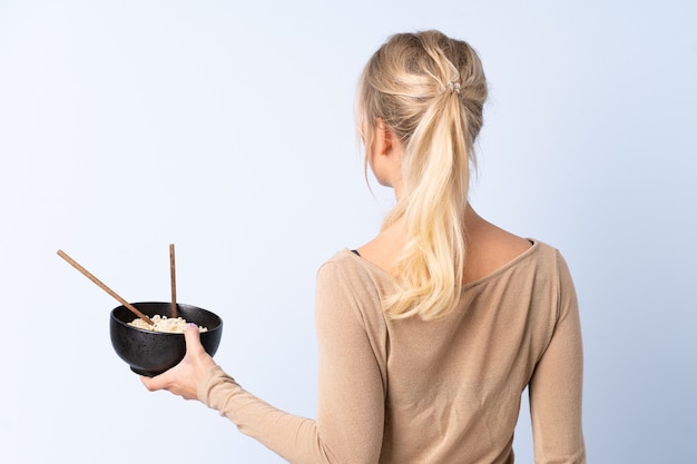 Femme blonde sur mur bleu isolé en position arrière tout en tenant un bol de nouilles avec des baguettes