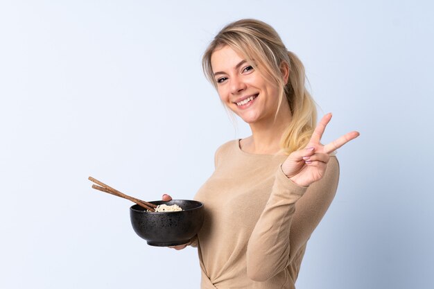 Femme blonde sur mur bleu isolé souriant et montrant le signe de la victoire tout en tenant un bol de nouilles avec des baguettes