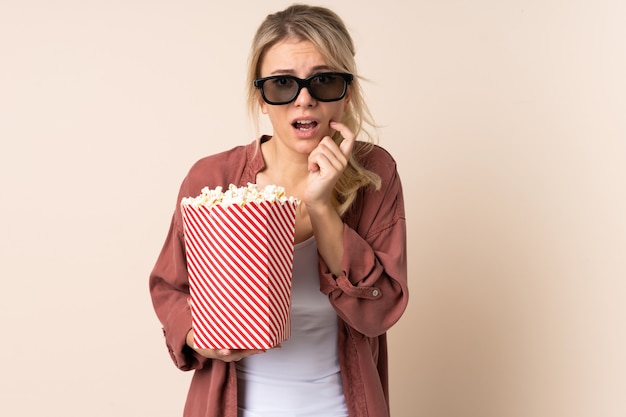 Femme blonde sur un mur isolé avec des lunettes 3D et tenant un grand seau de pop-corn