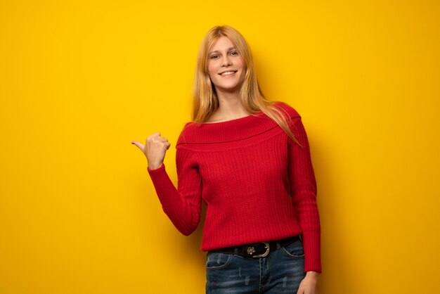 Femme blonde sur un mur jaune pointant sur le côté pour présenter un produit