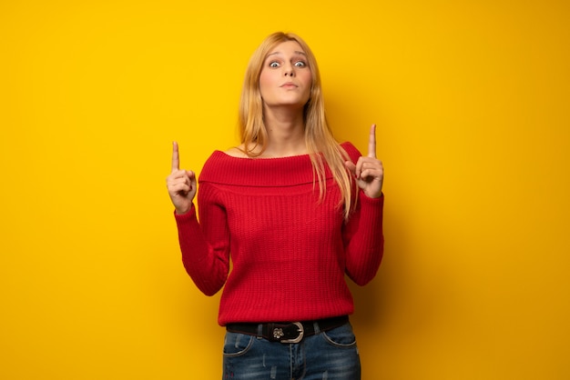 Femme blonde sur un mur jaune pointant vers le haut et surprise