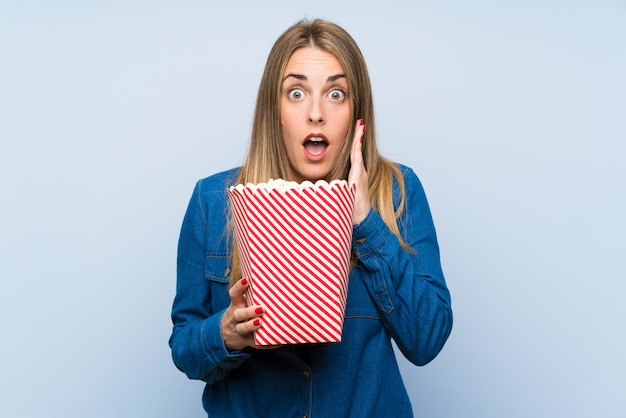 Femme blonde avec des pop-corn sur mur bleu faisant un geste de surprise