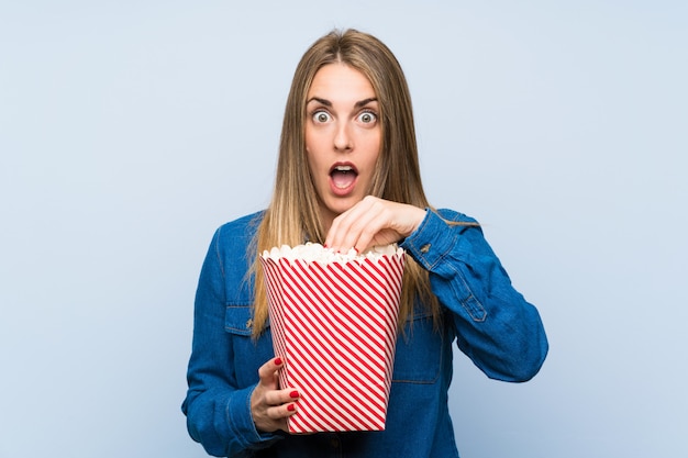 Femme blonde avec des pop-corn sur mur bleu faisant un geste de surprise