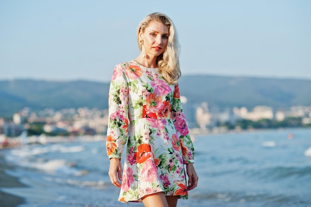 Femme blonde portant une robe debout sur une plage de sable au bord de la mer en profitant de la vue sur le coucher du soleil