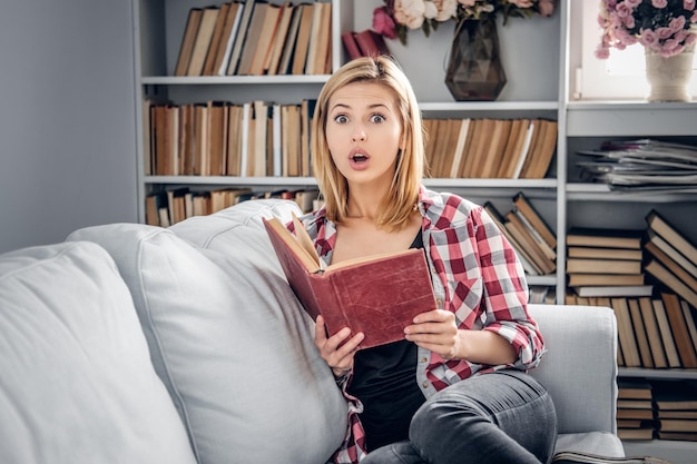 Une femme blonde positive est assise sur un canapé et lit un livre dans un salon.