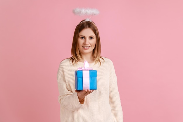 Femme blonde positive avec halo sur la tête tenant une boîte-cadeau enveloppée dans les mains donnant un cadeau