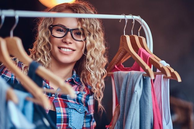 Une femme blonde positive à lunettes noires choisit des vêtements à la mode sur le portemanteau.