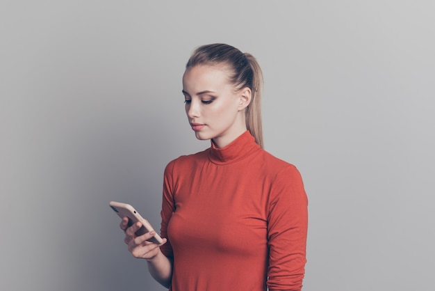 Femme blonde avec un pull à col roulé rouge posant contre le mur gris