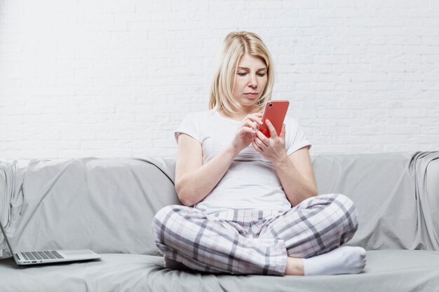 Une femme blonde en pyjama est assise sur un canapé gris avec un ordinateur portable et un téléphone. Travail et formation à distance pendant la pandémie de coronavirus.
