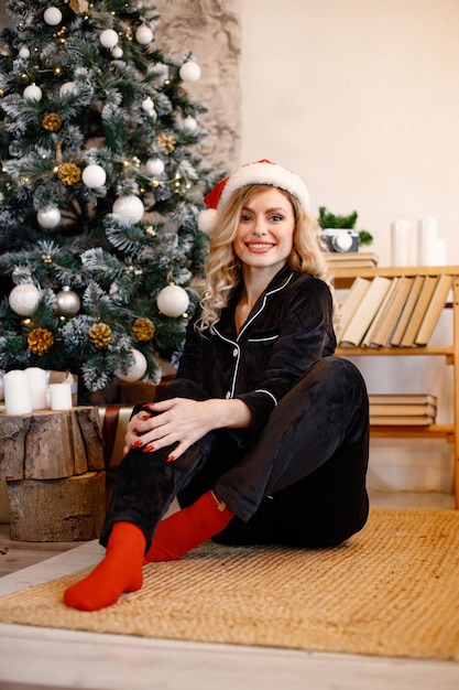 Une femme blonde en pyjama noir assise et posant près de l'arbre de Noël