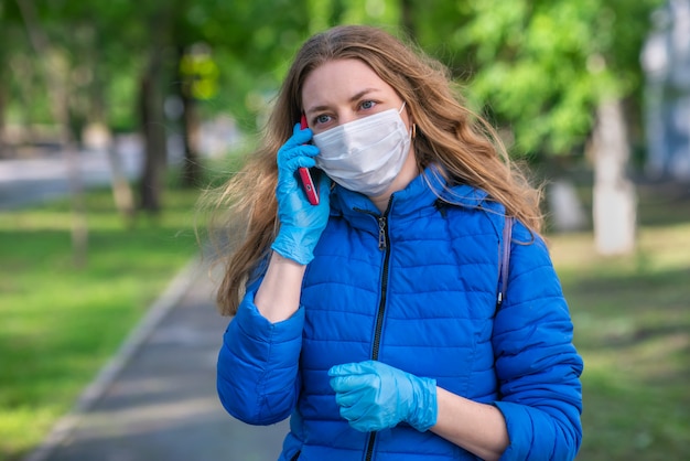 Une femme blonde de race blanche en masque de protection et des gants marche le long d'une rue vide et parle au téléphone. Comportement sûr