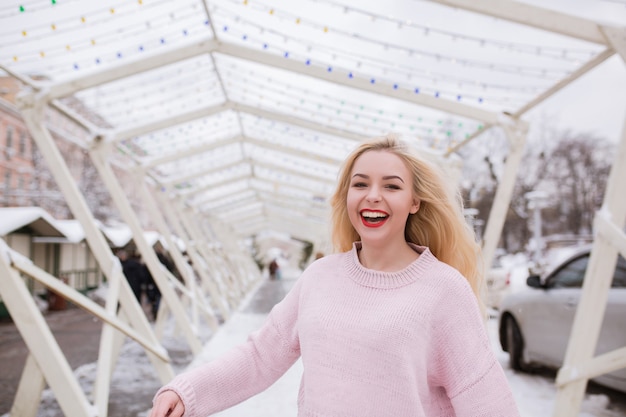 Femme blonde riante avec un maquillage lumineux, portant un pull rose, posant dans la rue en hiver