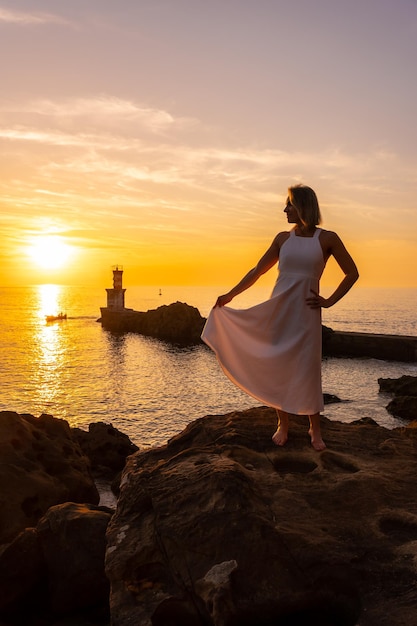 Une femme blonde en robe blanche au coucher du soleil à côté d'un phare dans la mer mode de vie sain et naturiste