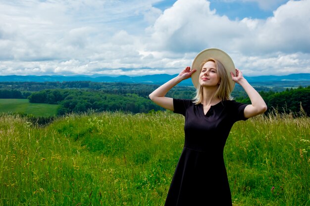 Femme blonde en robe noire et chapeau blanc au pré avec des montagnes