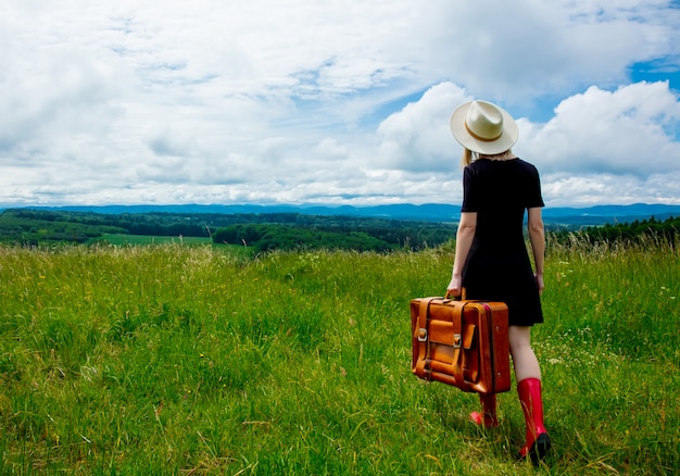 Femme blonde en robe noire et valise au pré avec des montagnes