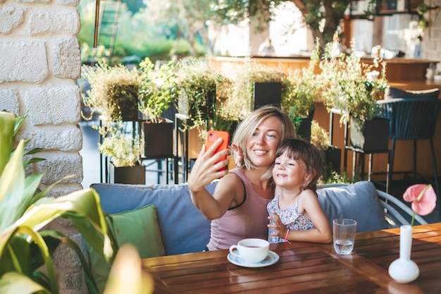 Femme blonde et sa petite fille utilisant un smartphone pour prendre un selfie dans le café