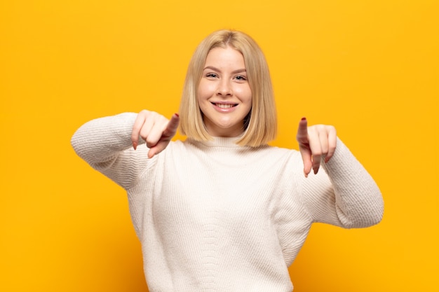 Femme blonde se sentant heureuse et confiante, pointant vers la caméra avec les deux mains et en riant, en vous choisissant