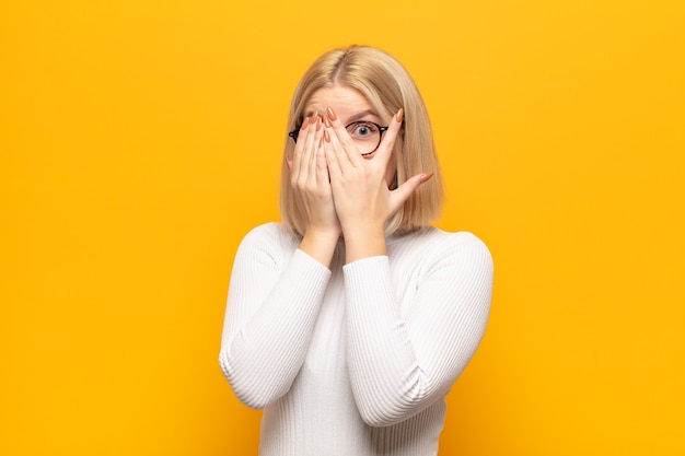 Femme blonde se sentir effrayée ou gênée, furtivement ou espionnage avec les yeux à moitié couverts de mains