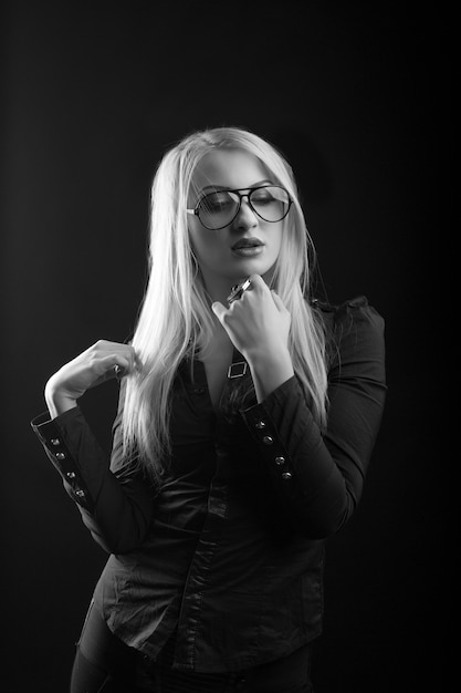 Une femme blonde séduisante aux cheveux longs porte des lunettes et un chemisier posant au studio. Prise de vue monochrome