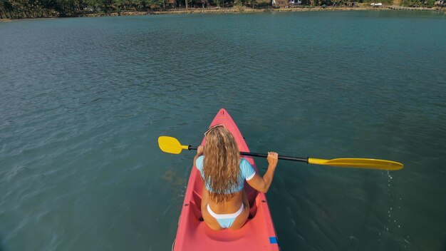 Une femme blonde séduisante en maillot de bain bleu navigue sur un kayak en plastique rose le long de la mer d'azur à l'arrière de la station balnéaire exotique en haut au-dessus de la vue. Voyager dans les pays tropicaux. La fille navigue sur le kayak dans l'océan.