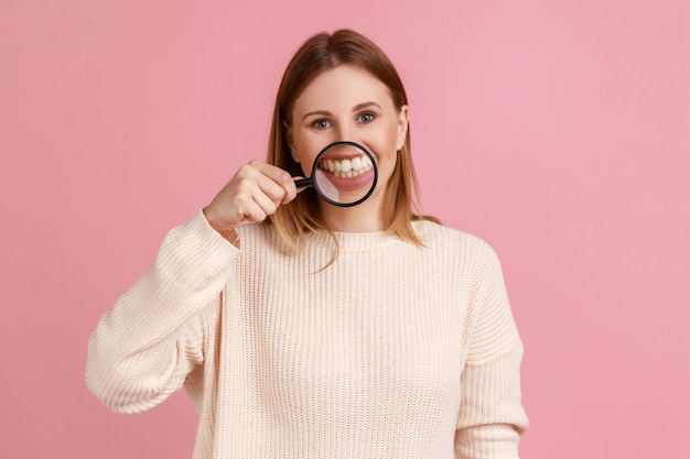 Femme blonde séduisante positive debout tenant une loupe sur ses dents en regardant la caméra