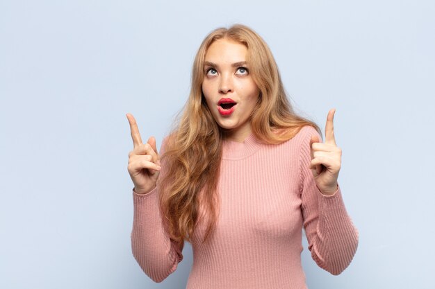 Femme blonde semblant choquée, étonnée et bouche ouverte, pointant vers le haut avec les deux mains pour copier l'espace