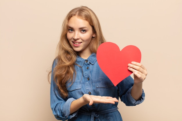 Femme blonde semblant surprise et choquée, avec la mâchoire tombée tenant un objet avec une main ouverte sur le côté