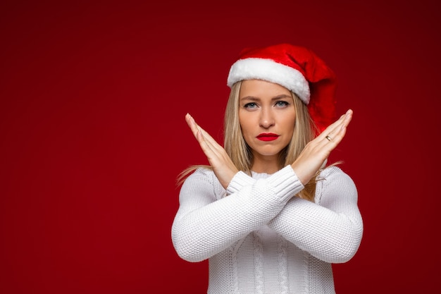 femme blonde sérieuse en bonnet de Noel et pull blanc montrant les bras croisés. Concept d'interdiction.