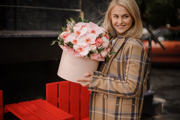 Femme blonde souriante en manteau d'automne à carreaux avec une boîte rose de fleurs