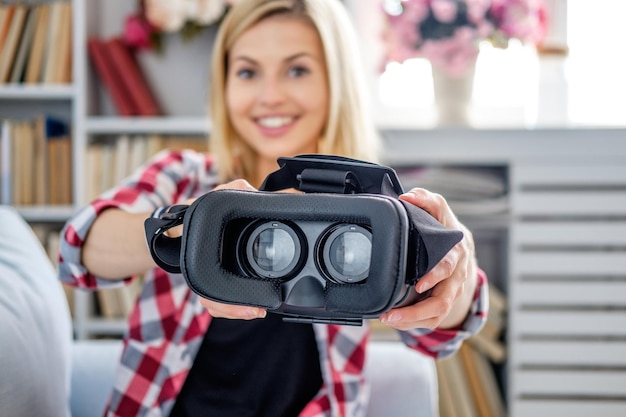 Femme blonde souriante montrant des lunettes de réalité virtuelle dans un salon.